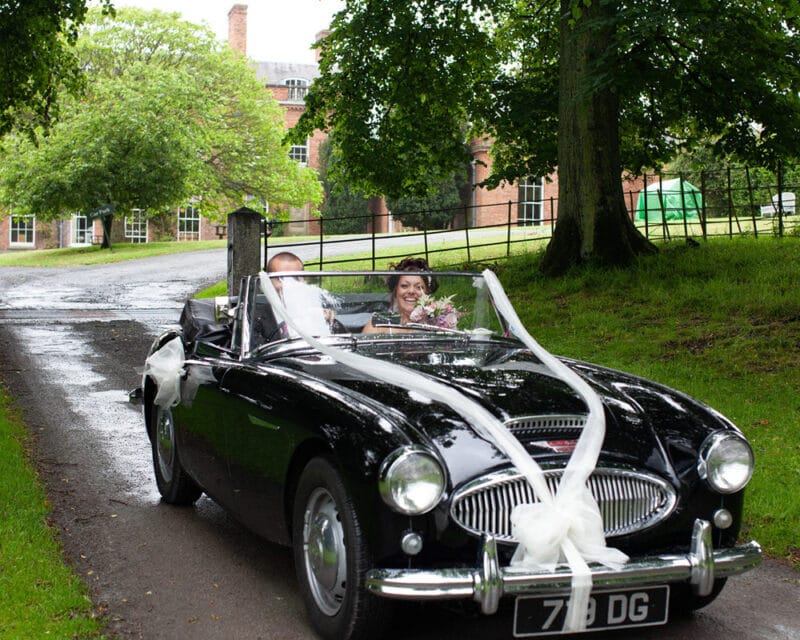 Bride smiling in a black vintage wedding car driving through a countryside venue entrance