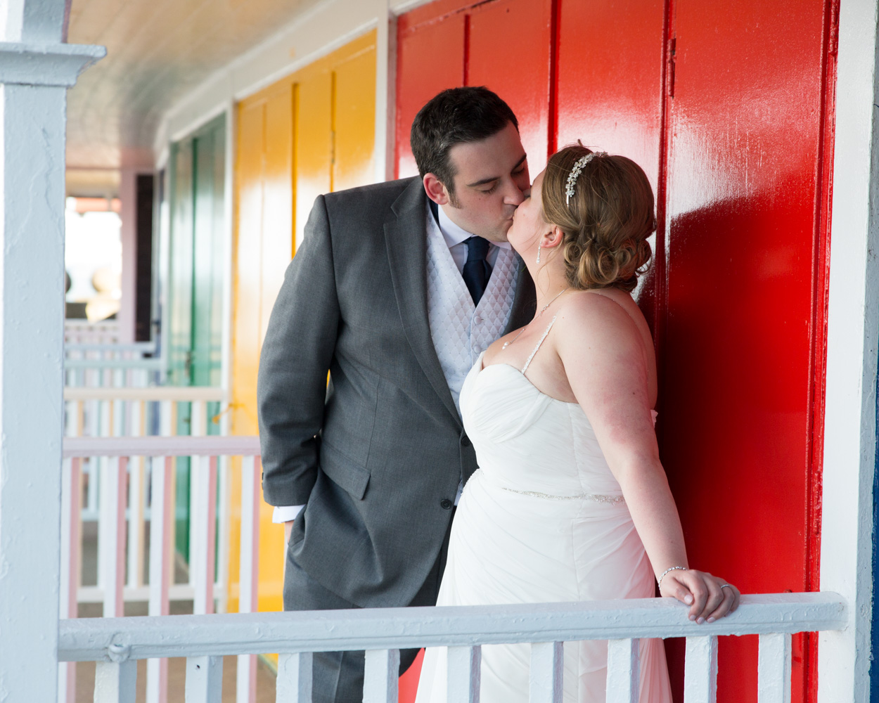Lisa & Oliver standing by the colourful beach cabins at Scarborough Spa, May wedding couple portrait, smiling in the wind.