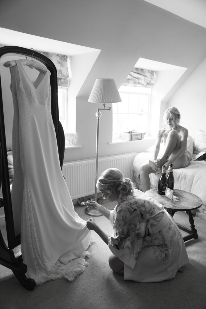 Bride sat on the edge of the bed - her sister helping sew the bottom edge of the wedding dress - black and white documentary photography