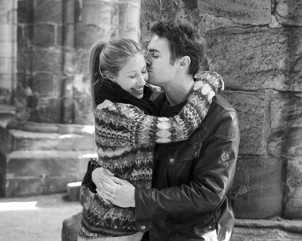 Andrew kisses Zoe’s cheek as she smiles, seated on a weathered stone column inside Kirkstall Abbey.