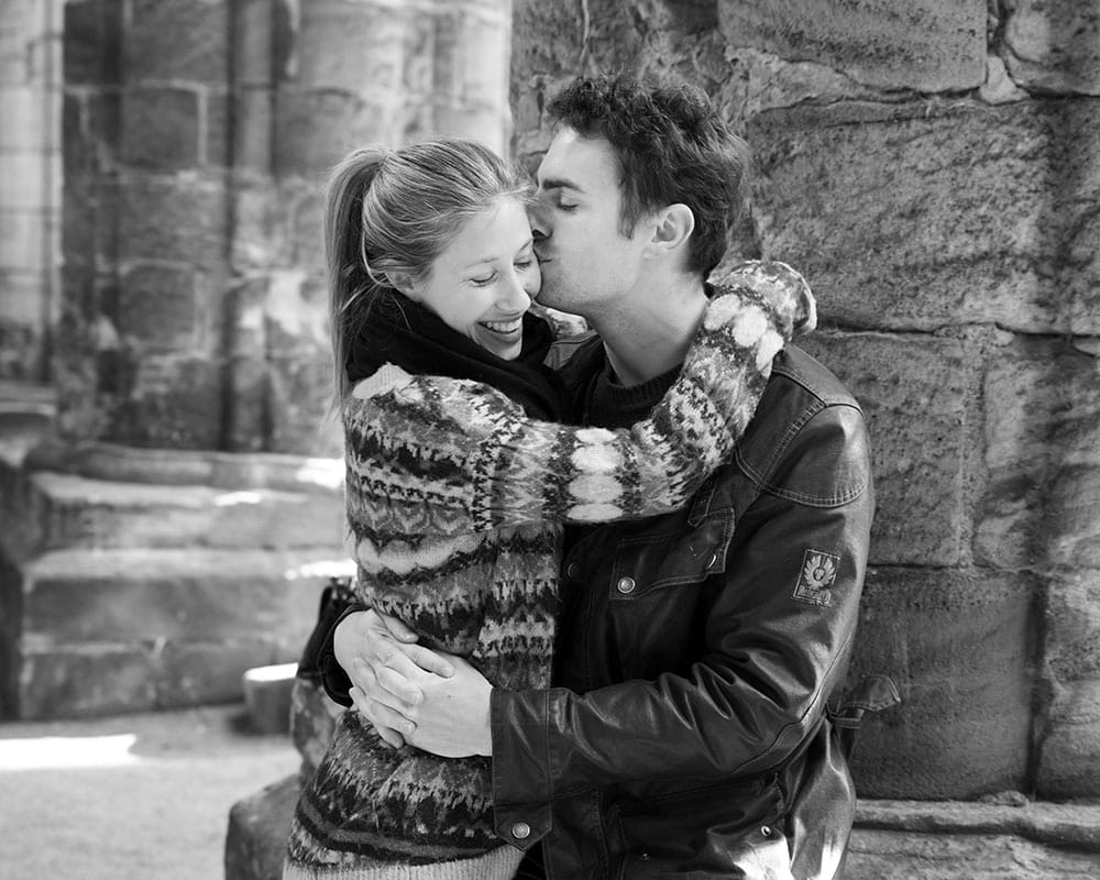 Andrew kisses Zoe’s cheek as she smiles, seated on a weathered stone column inside Kirkstall Abbey.