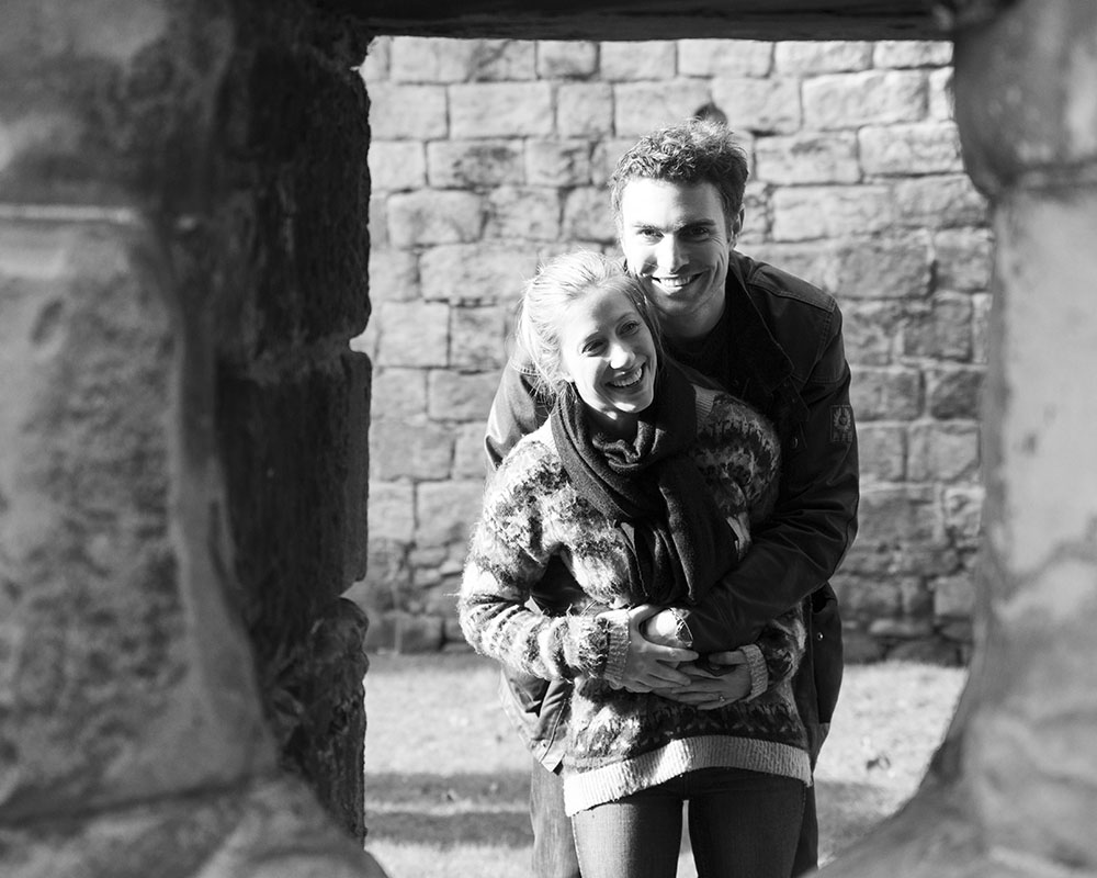 Couple hugging and peeking playfully through a carved stone window at Kirkstall Abbey.