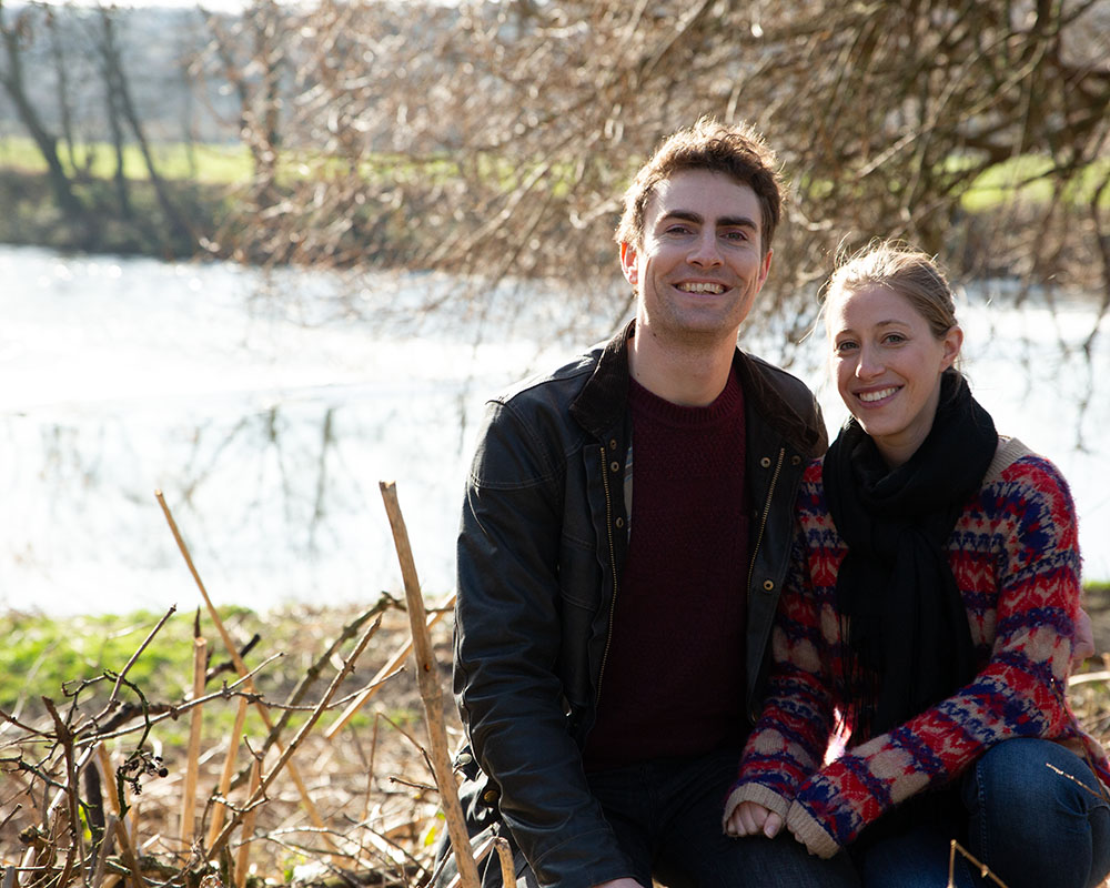 Close portrait of the couple by the river at Kirkstall Abbey, smiles soft and sleeves pulled over chilled hands.