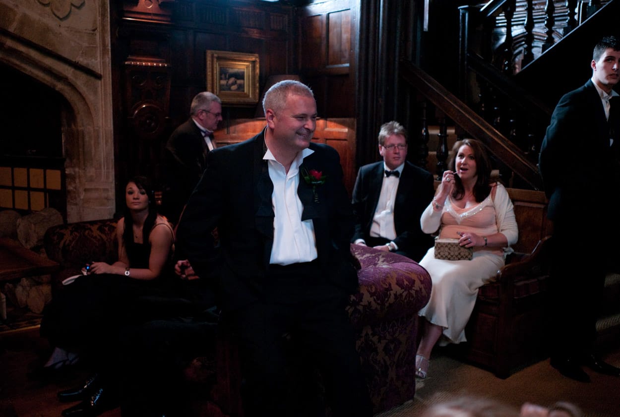 Groom smiling while seated on a sofa surrounded by guests.