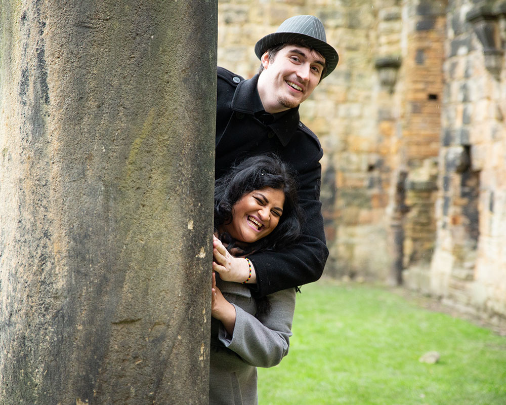 Zehra and Richard laughing as they peek from behind a stone column at Kirkstall Abbey in Leeds.