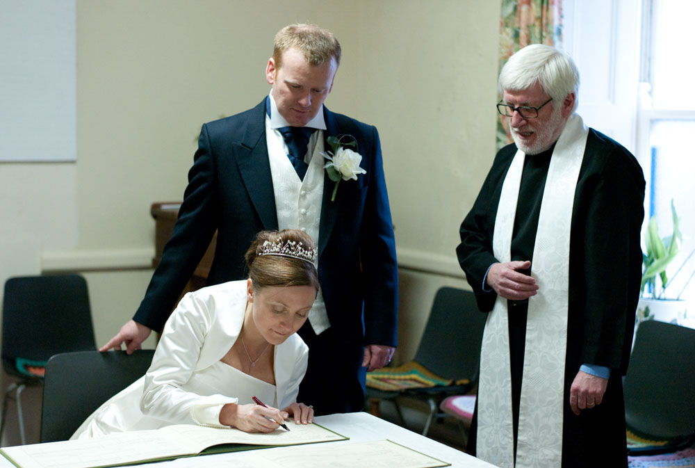The bride signs the register as the groom and minister look on.