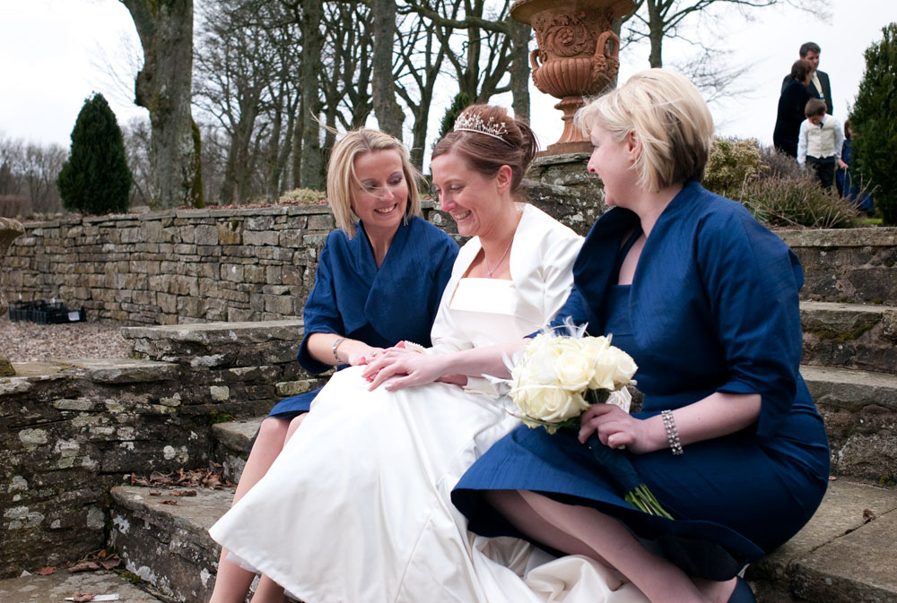 The bride and her two bridesmaids in blue dresses sit smiling on the cold stone steps.