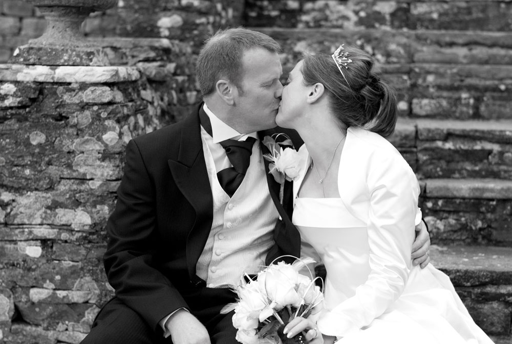 The couple sit together on the steps, sharing a kiss in black and white.