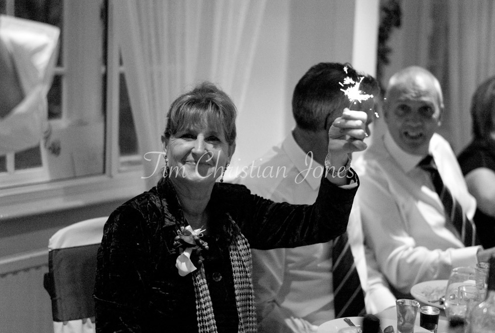 The bride’s mum lifts her sparkler with joy, light dancing across her face.