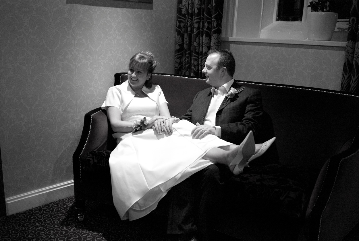 Bride resting legs across groom’s legs on sofa in black and white