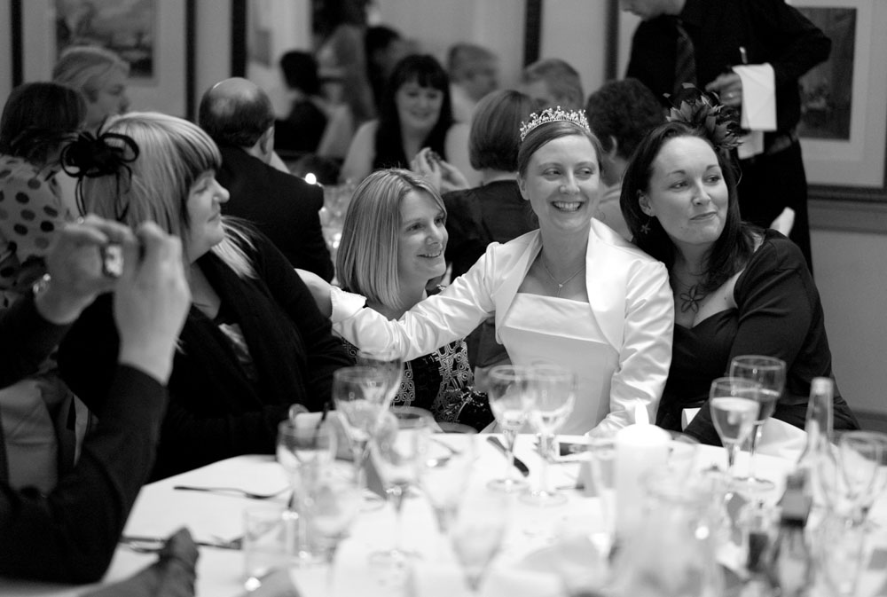 Black and white shot of the bride and three friends posing while others at the table watch on.