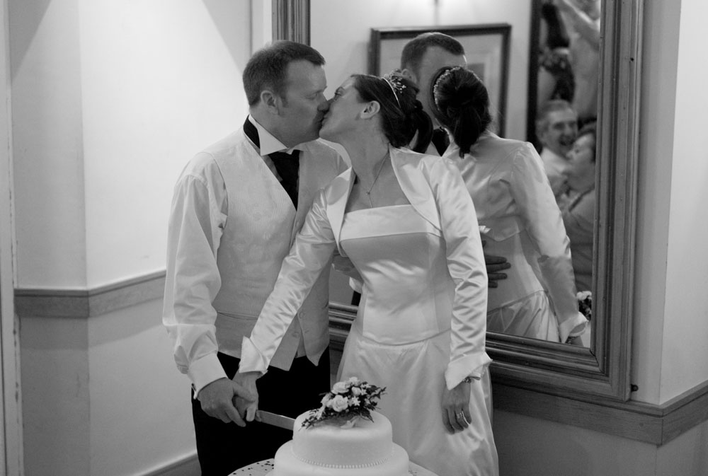 Black and white photo of the couple kissing as they cut the cake together.