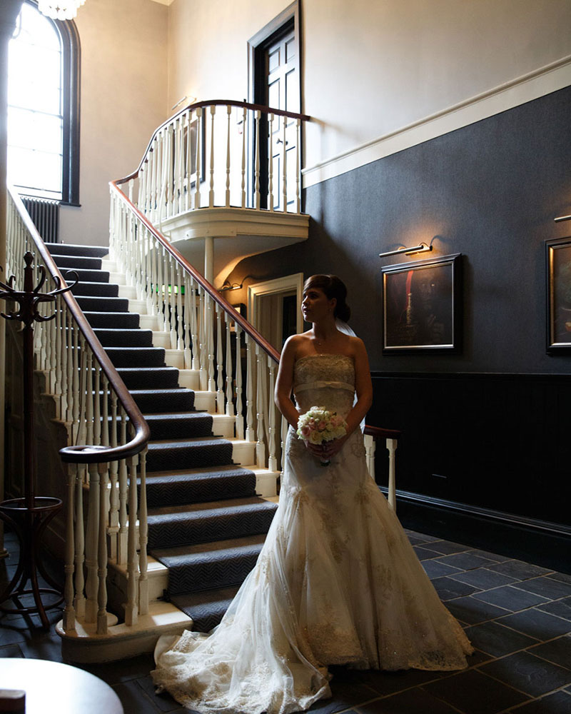 Bride in wedding dress at Grays Court in York, standing at the foot of a staircase with soft window light catching her silhouette and flowing dress train