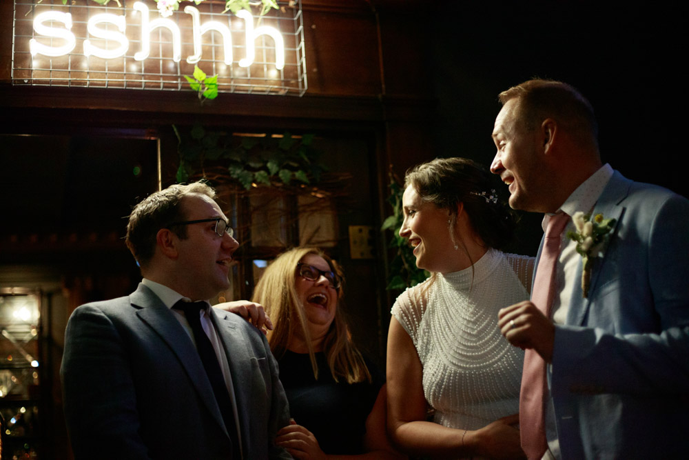 Gemma and Danny with Best man and wife smiling under the sshhh neon sign above the entrance to the evening reception room - lost and found Leeds club.