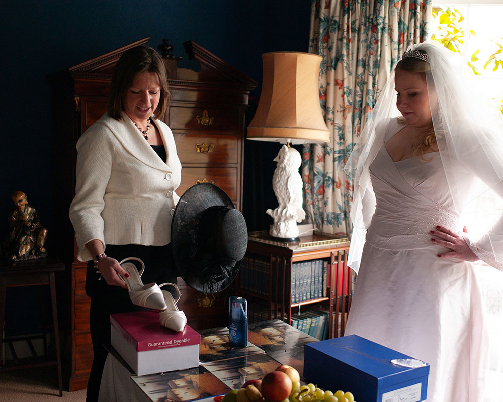 Bride’s wedding shoes being lifted from a box by her friend during morning wedding preparations, with soft natural light filling the room as the bride looks on.