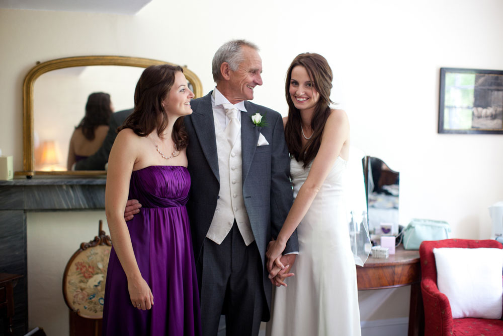 Natural wedding preparation moment with the bride, her father, and sister smiling together—she gently holds her father’s arm while they share an emotional, candid moment before the ceremony