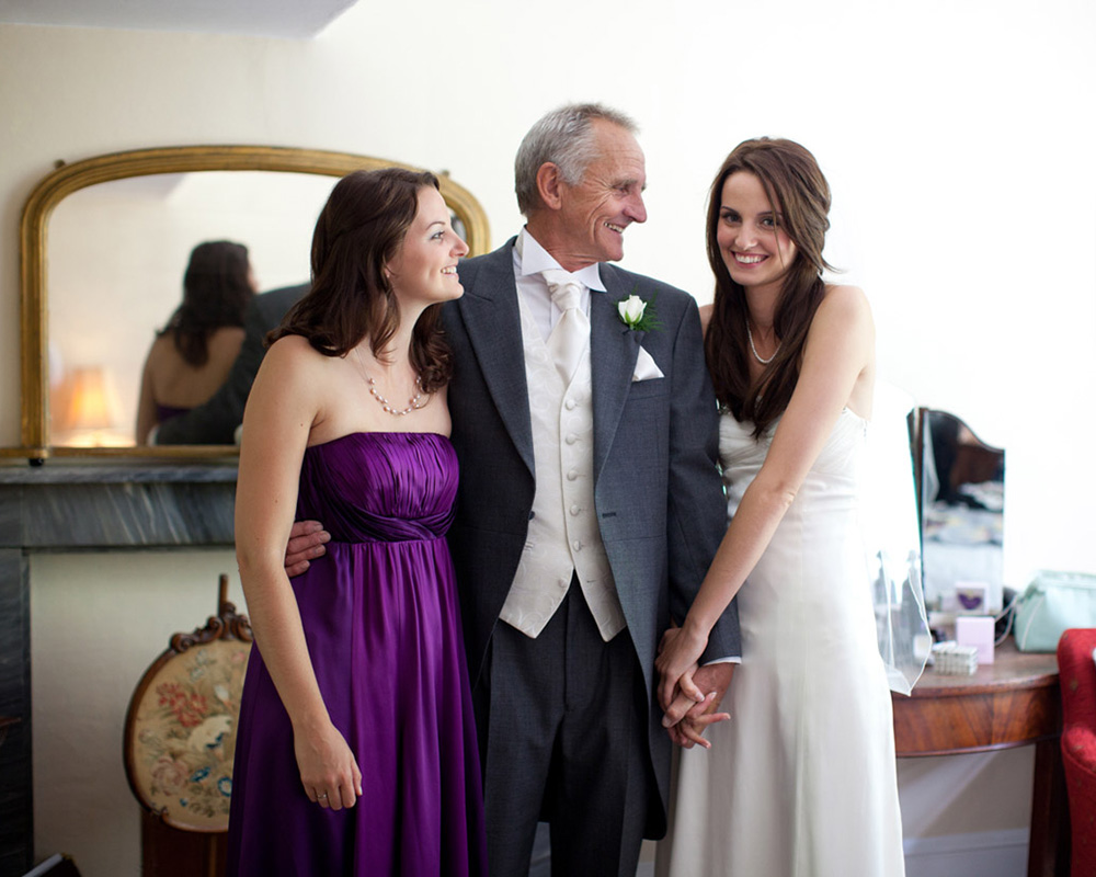 Natural wedding preparation moment with the bride, her father, and sister smiling together—she gently holds her father’s arm while they share an emotional, candid moment before the ceremony