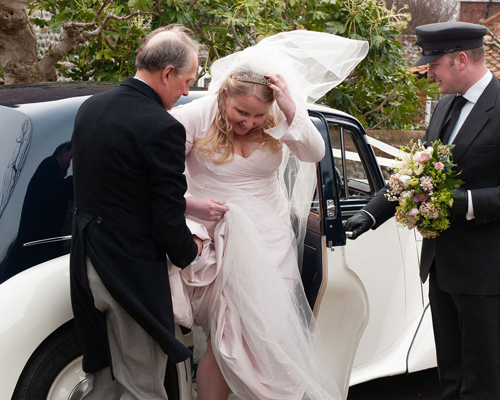 Bride stepping from a vintage car as wind lifts her veil, smiling on arrival.