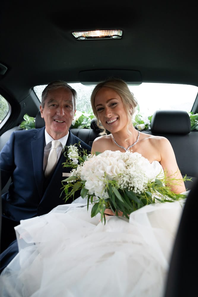 Bride and her father share a smile in the wedding car, bouquet of white flowers and ferns.