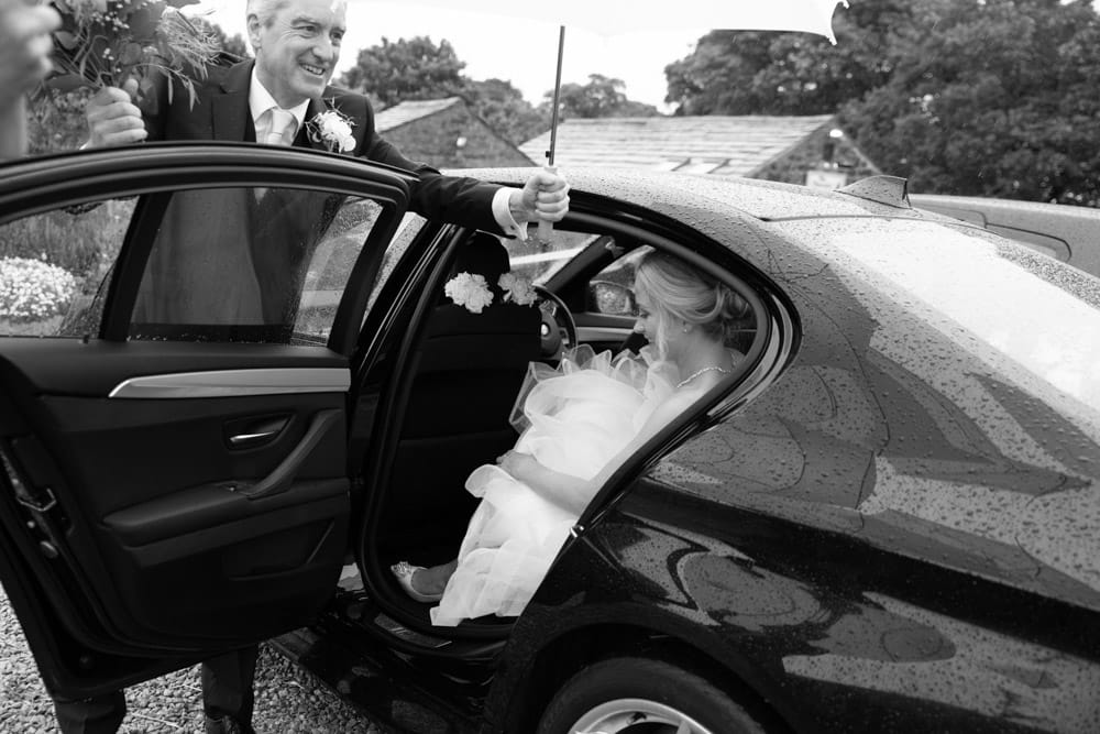 Father of the bride holds an umbrella to protect his daughter about to enter the wedding venue Chilli Barn Otley.