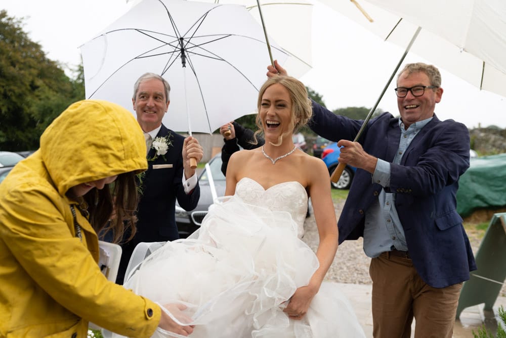 Bride laughs as bridesmaids and staff hold umbrellas.