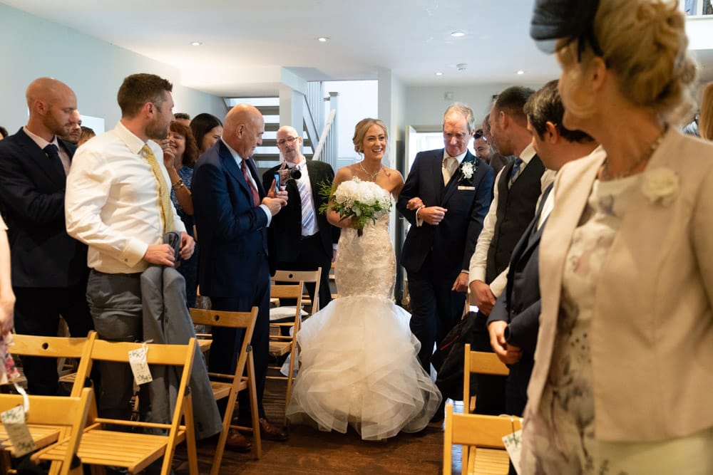 Bride in lace smiles at guests as she walks down the aisle with her father.