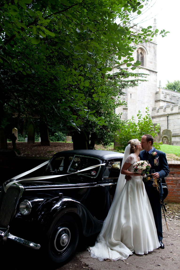 Couple kiss - flowing white dress - military uniform - stood next to black vintage wedding car - the church softly in the background.