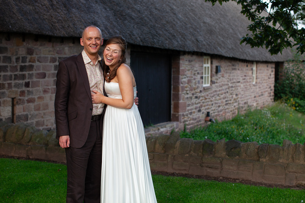 Village hall wedding reception couple portrait on a grass bank beside a stone and thatched village hall in Yorkshire at dusk—the bride laughing and the groom smiling in a natural, relaxed moment captured by Leeds wedding photographer Tim Christian Jones