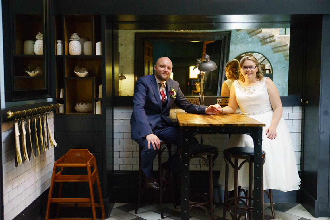 Couple portrait inside the Churchill Hotel in York - sat at a bar table - holding hands and smiling for the camera.