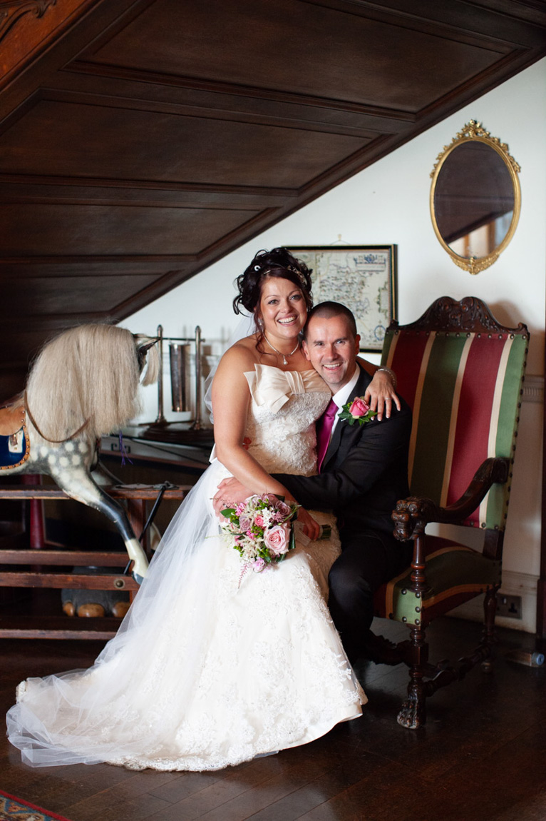 Couple inside an historic wedding venue - Ella and sat on an old chair - a vintage rocking horse and details. They smile naturally captured by a Leeds wedding photographer.