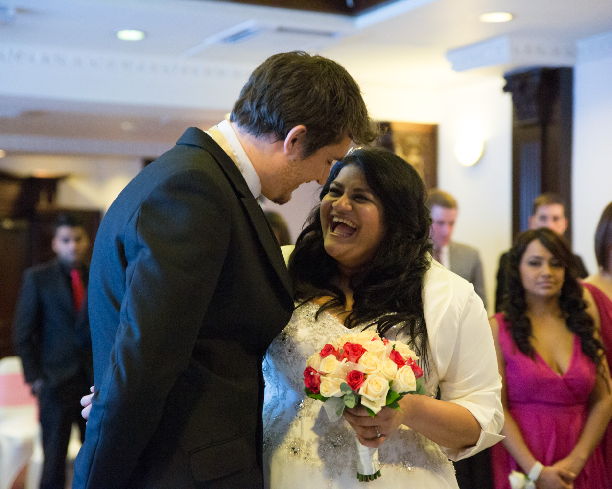 Groom playfully pretends to sign the register, bride looks at guests laughing