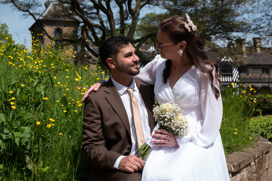 Colourful couple portrait - Inga sat on Roland's lap - sat on a stone wall amidst green and buttercups - Shibden Hall in the background. By Tim Christian Jones Photography | Leeds wedding photographer.