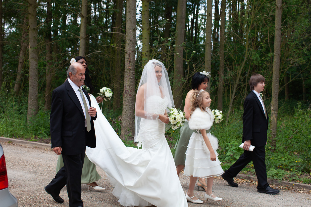 Bride walks to the ceremony with her bridal party, train held by her friend.