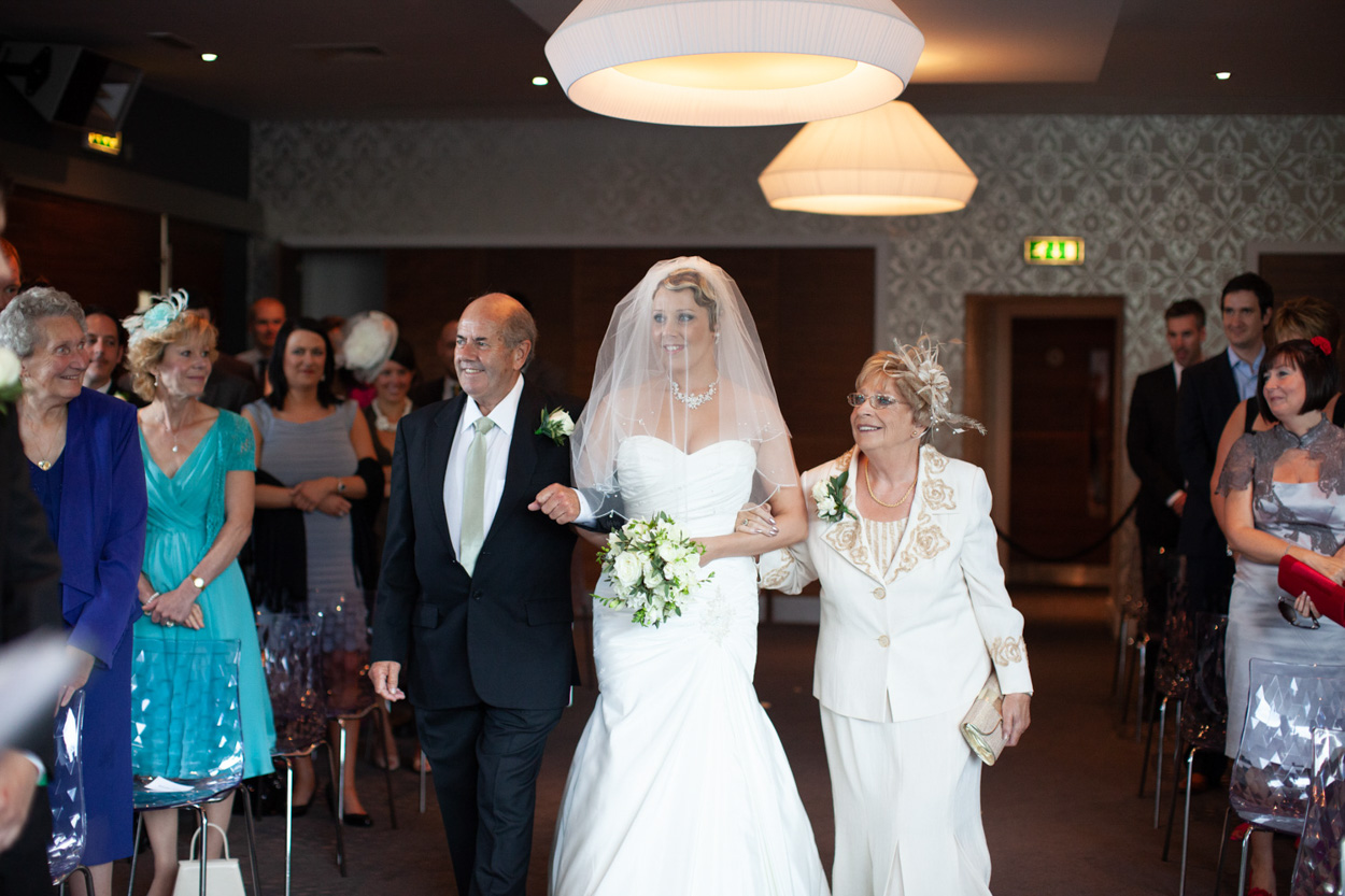 Bride walks in with her parents, one on each arm, beaming with pride.