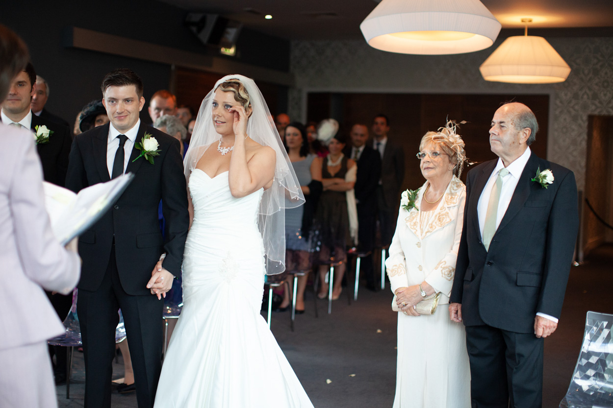 Groom smiling, bride wiping away a tear as parents stand nearby.