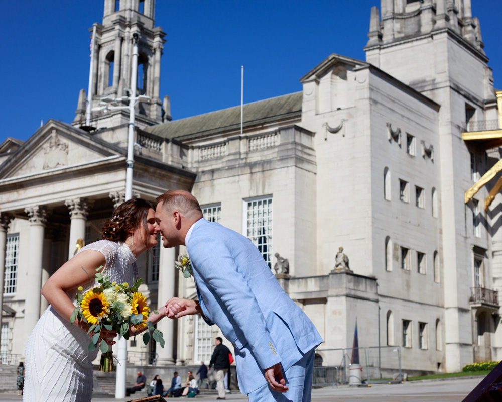 Couple portrait of Gemma and Danny sharing a playful moment beside piano festival art on Millennium Square in Leeds, with Leeds Civic Hall in the background on a bright blue sky wedding day.