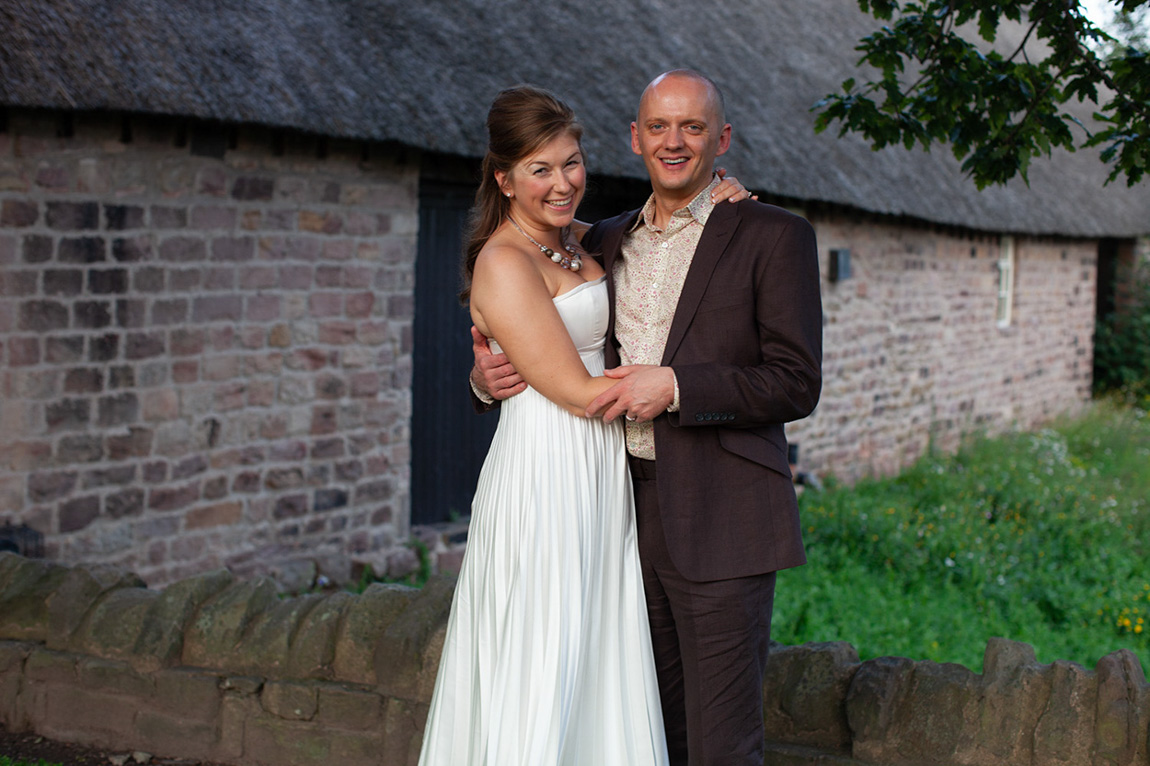 Village hall wedding reception couple portrait on a grass bank beside a stone and thatched village hall in Yorkshire at dusk—the bride laughing and the groom smiling in a natural, relaxed moment captured by Leeds wedding photographer Tim Christian Jones