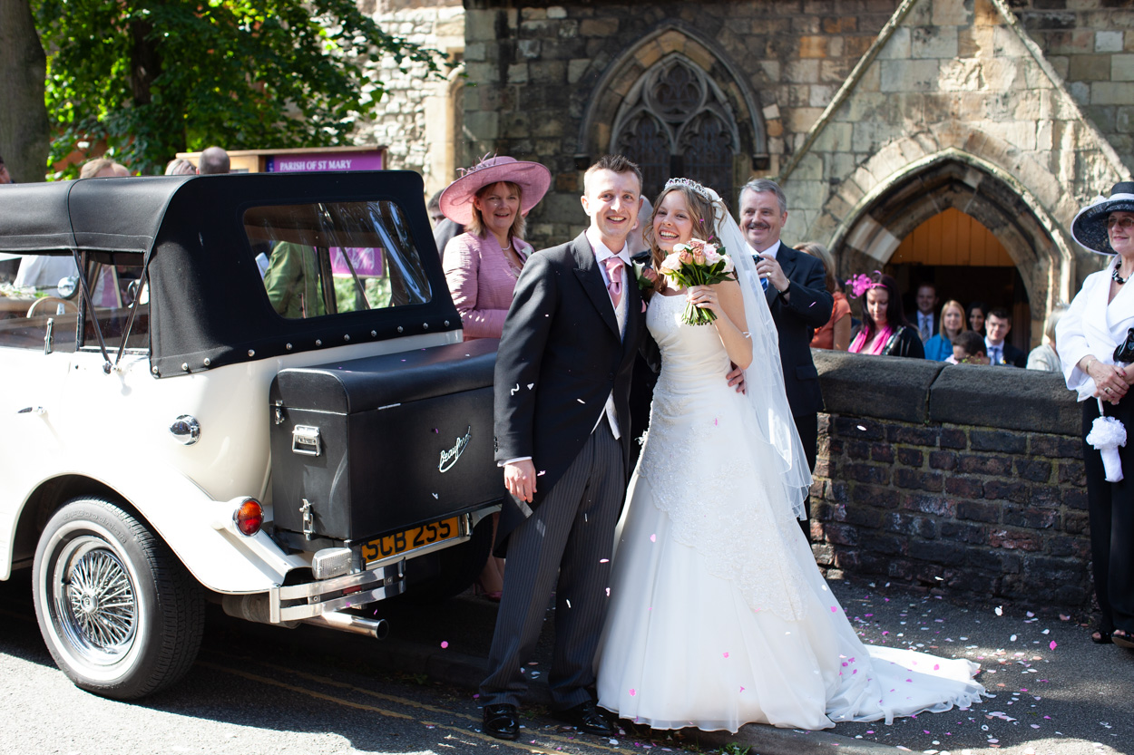 Bride and groom standing by their wedding car outside the church, guests and family gathered around in a natural moment of celebration - photographed near Leeds.
