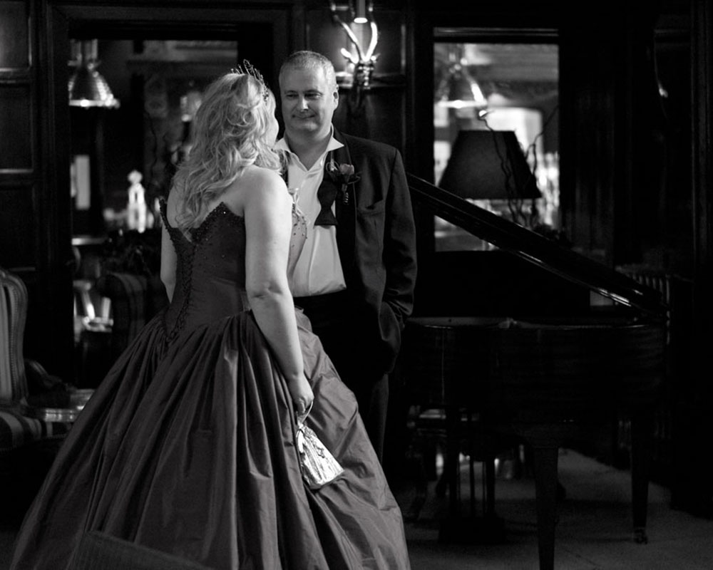 Couple standing quietly beside a piano before entering their wedding breakfast, captured in black and white.