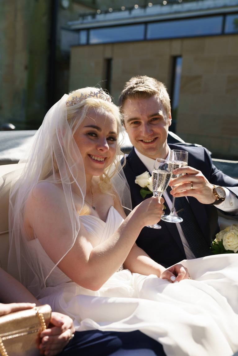 Holly and James clink champagne glasses sat in the vintage wedding car outside The Mansion in Leeds — captured naturally by Leeds wedding photographer Tim Christian Jones.