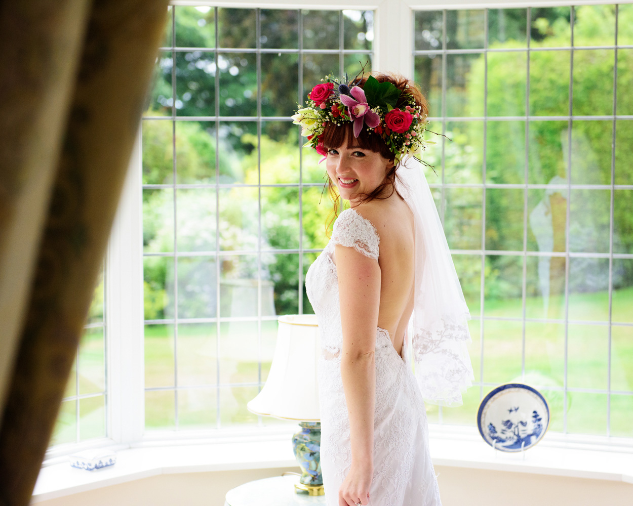 Bride stands by the window with floral crown and veil — wedding morning portrait.