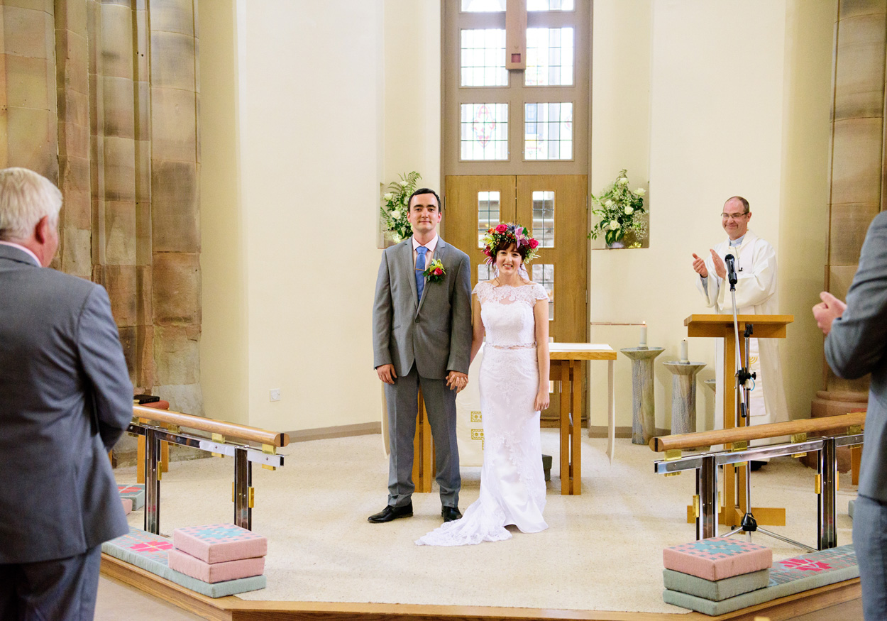 Groom lifts the bride playfully beneath an arch, church behind, bride points her ballet-style shoes.