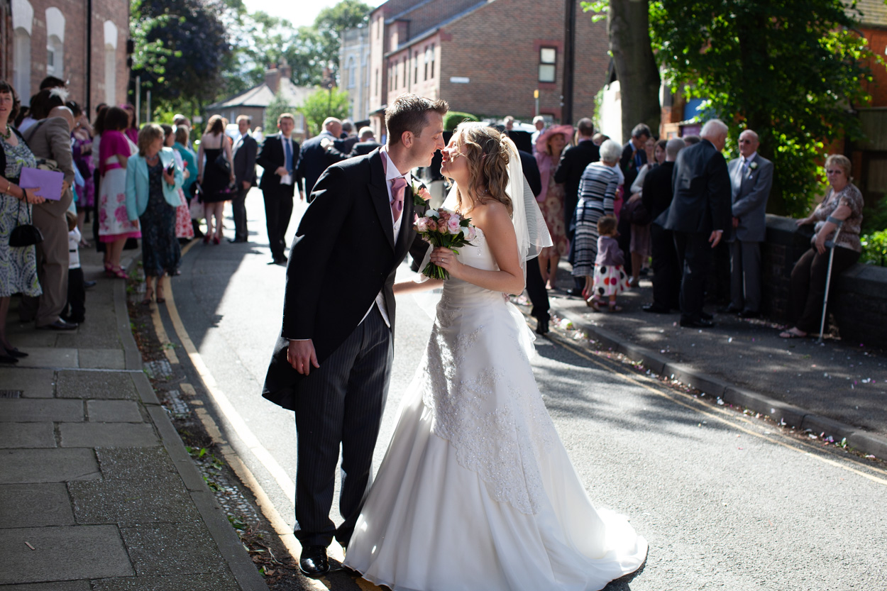 Bride and groom standing in sunlight about to kiss, guests and strangers lining the road behind in a lively wedding scene