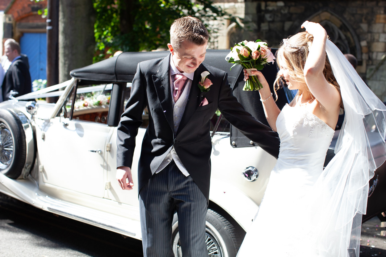 Groom gently helping the bride step into the road while she holds her veil, guests and onlookers gathered behind