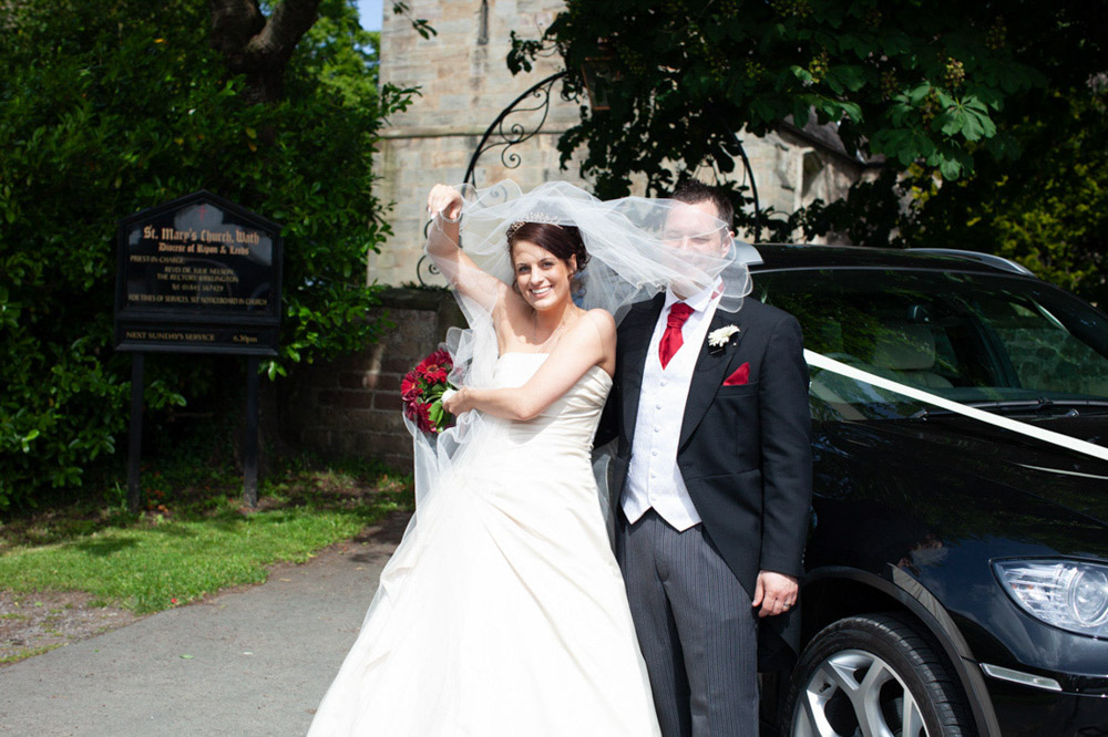 Bride and groom laughing as the wind lifts her veil by their wedding car at a wrought iron arch entrance to the church pathway at St Mary’s Church in Wath—natural candid wedding moment captured by Tim Christian Jones