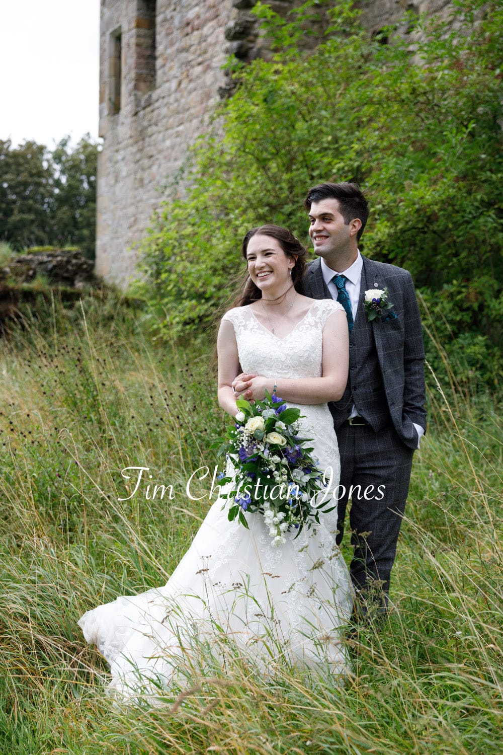 Couple stood together in long grass in the Yorkshire Dales, hands meet, relaxed and smiling.